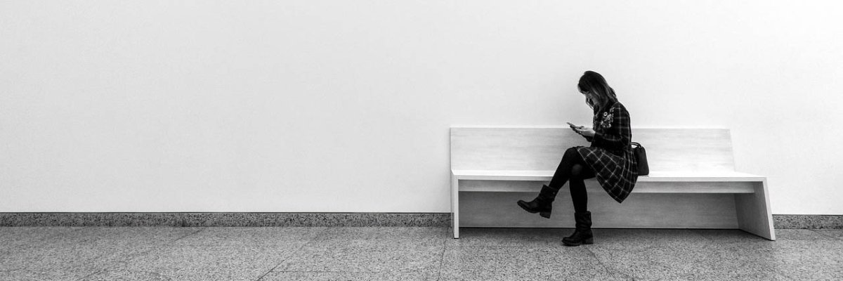 Eine Person sitzt auf einer Bank und schaut auf ihr Handy vor einer weißen Wand