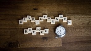 Auf einer Holzplatte liegt der Schriftzug Einrichtung buchen und eine Taschenuhr Auf einer Holzplatte liegt der Schriftzug Einrichtung buchen und eine Taschenuhr